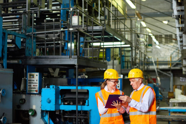 Kvinne og mann under en sikkerhetsinspeksjon på en fabrikk, iført gule hjelmer og oransje sikkerhetsvester.